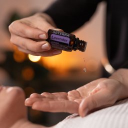 Karolina Ziemniak - Krople olejku lawendowego z buteleczki DoTerra spadają na dłoń osoby leżącej na białym ręczniku, w tle rozmyte światła.