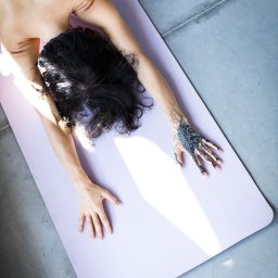 Karolina Ziemniak - Kobieta leży na plecach na fioletowej macie do ćwiczeń, ramiona wyciągnięte do przodu, dłoń z tatuażem skierowana w górę, widok z góry.