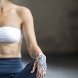 Karolina Ziemniak - Kobieta w pozycji lotosu z henną na dłoni, ubrana w sportowy strój, medytuje na tle szarej ściany.