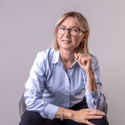 Karolina Ziemniak - Portret kobiety w okularach, siedzącej na krześle, w błękitnej koszuli i czarnych spodniach. Uśmiechnięta, z dłonią przy twarzy. Stonowane tło, naturalne światło.