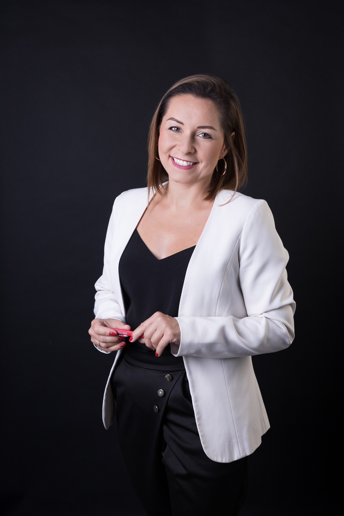 Uśmiechnięta kobieta w białej marynarce na czarnym tle, trzymająca w dłoniach różową szminkę, z widocznymi złotymi kolczykami kołami.