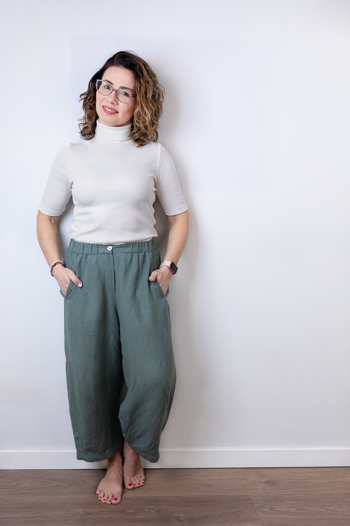 Uśmiechnięta kobieta w okularach, białym golfie i szerokich, zielonych spodniach z lnu, pozuje na tle białej ściany, z rękami w kieszeniach i bosymi stopami na drewnianej podłodze.