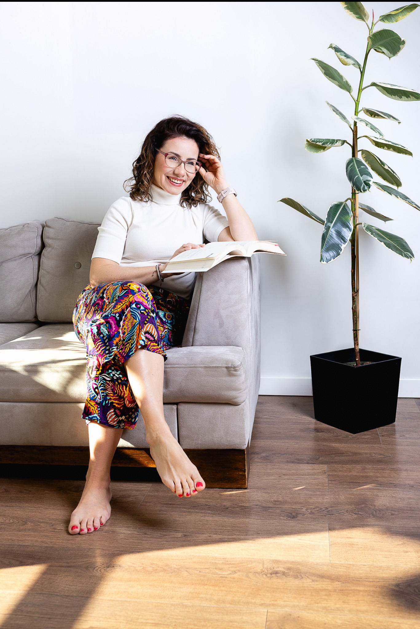 Kobieta w okularach, siedząca na szarej sofie w Tychach, czyta książkę w jasnym wnętrzu z rośliną doniczkową obok.