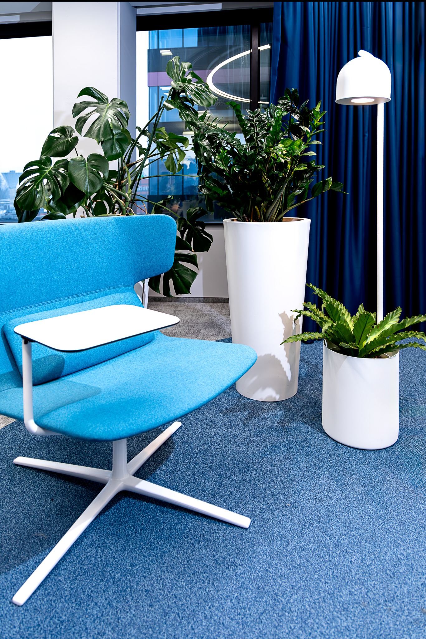 Nowoczesne biuro: błękitny fotel z białym blatem, białe donice z zielonymi roślinami, lampa podłogowa i zasłony na tle okna z widokiem na miasto.
