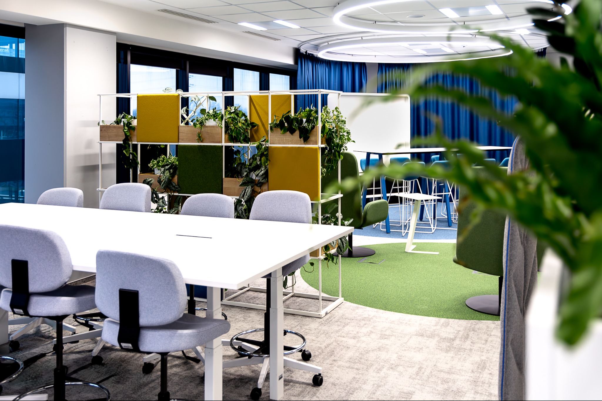 Nowoczesne biuro z białym stołem konferencyjnym, szarymi krzesłami na kółkach, modułową ścianką działową z roślinami i kolorowymi panelami, oraz dywanem w odcieniach szarości i zieleni.