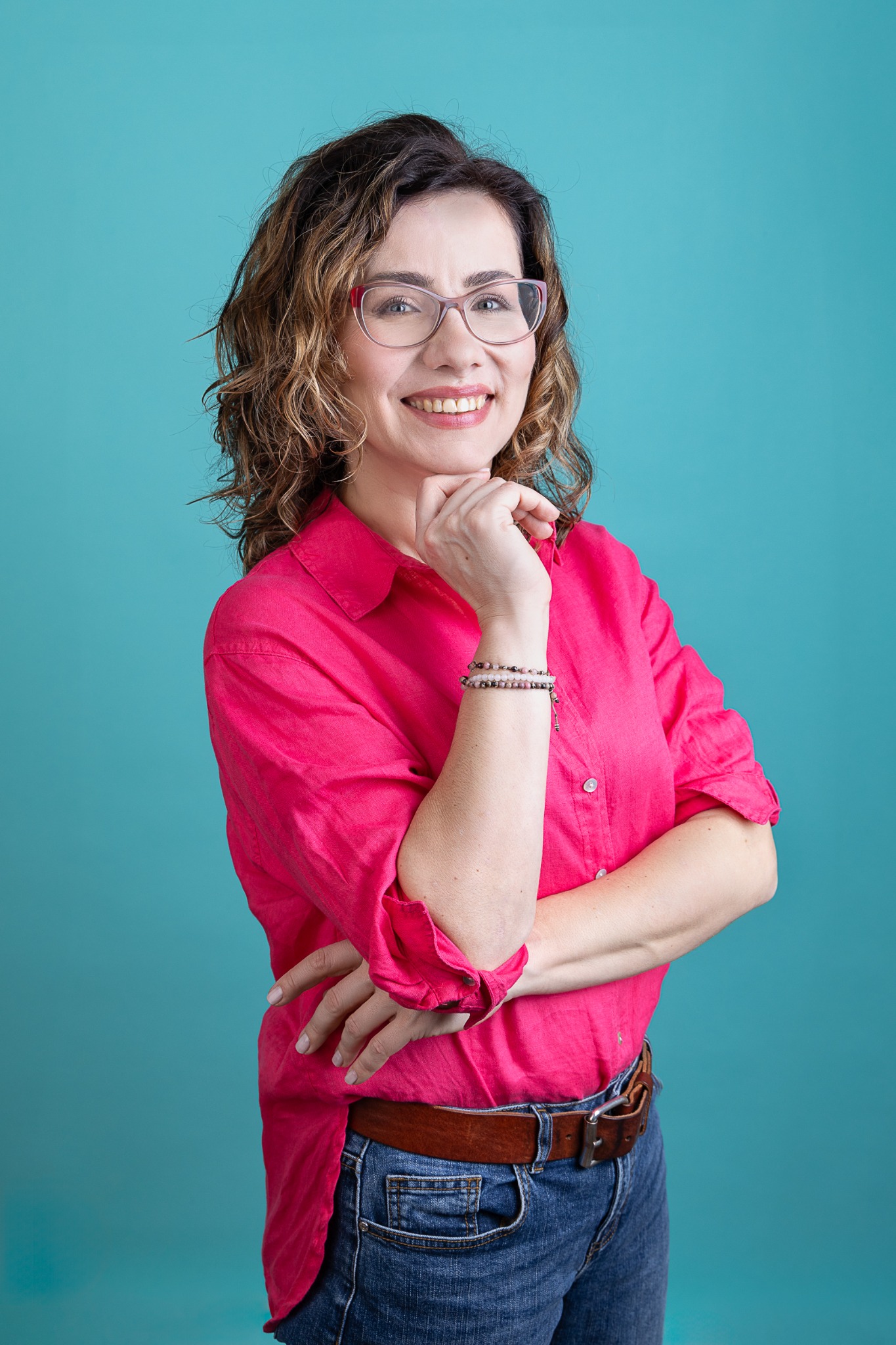 Portret uśmiechniętej kobiety w okularach z brązowymi kręconymi włosami, ubrana w różową koszulę, dżinsy i brązowy pasek, pozująca na tle turkusowego tła.