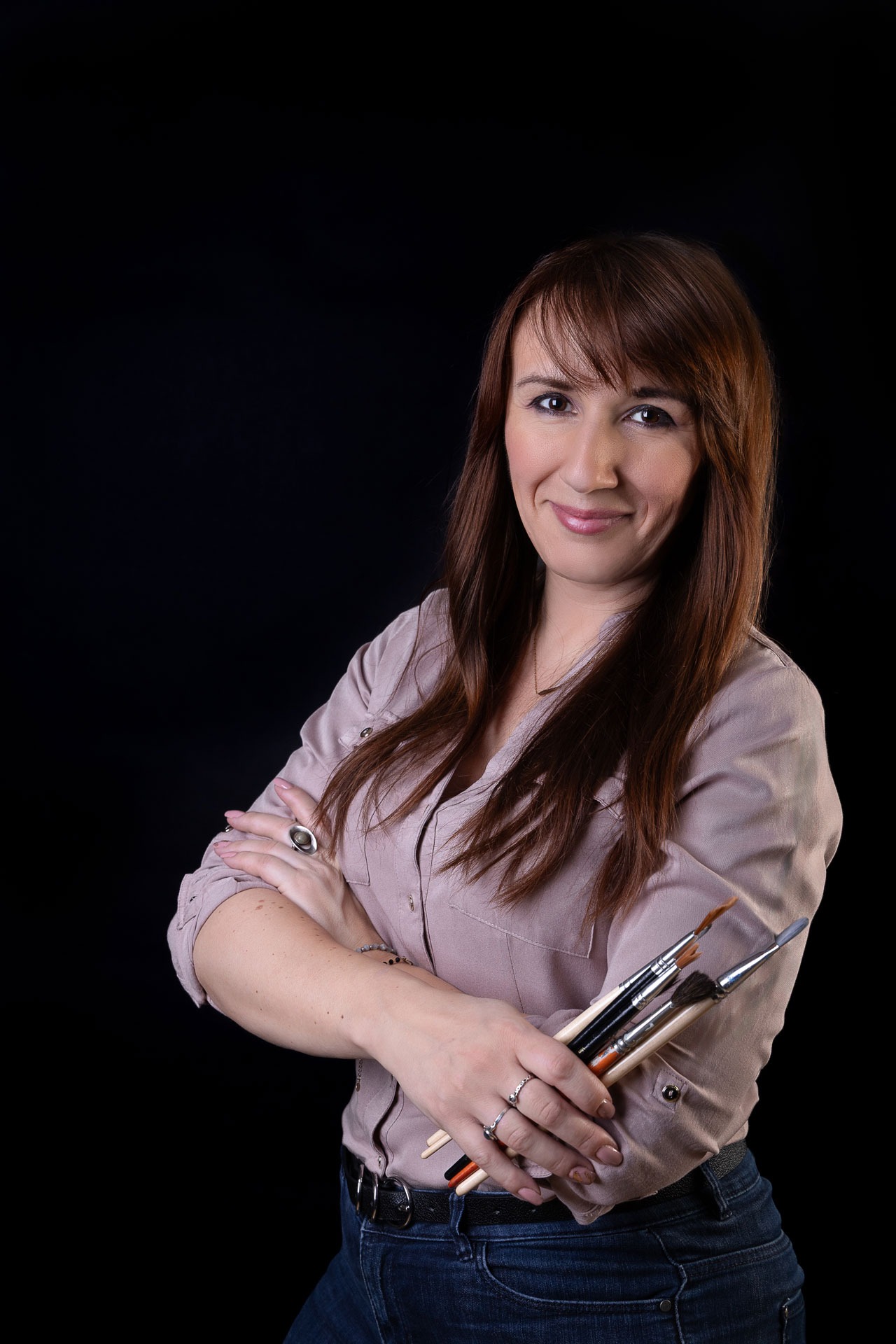 Portret kobiety z brązowymi włosami, trzymającej pędzle do makijażu na czarnym tle. Ubrana w dżinsy i koszulę, z pierścionkami na palcach.