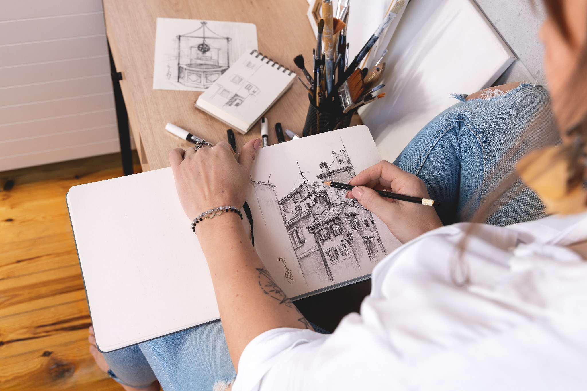 Osoba rysuje ołówkiem architekturę miejską w szkicowniku. Na drewnianym stole akcesoria do rysowania, pędzle i szkice. Ujęcie z góry, skupione na procesie twórczym.