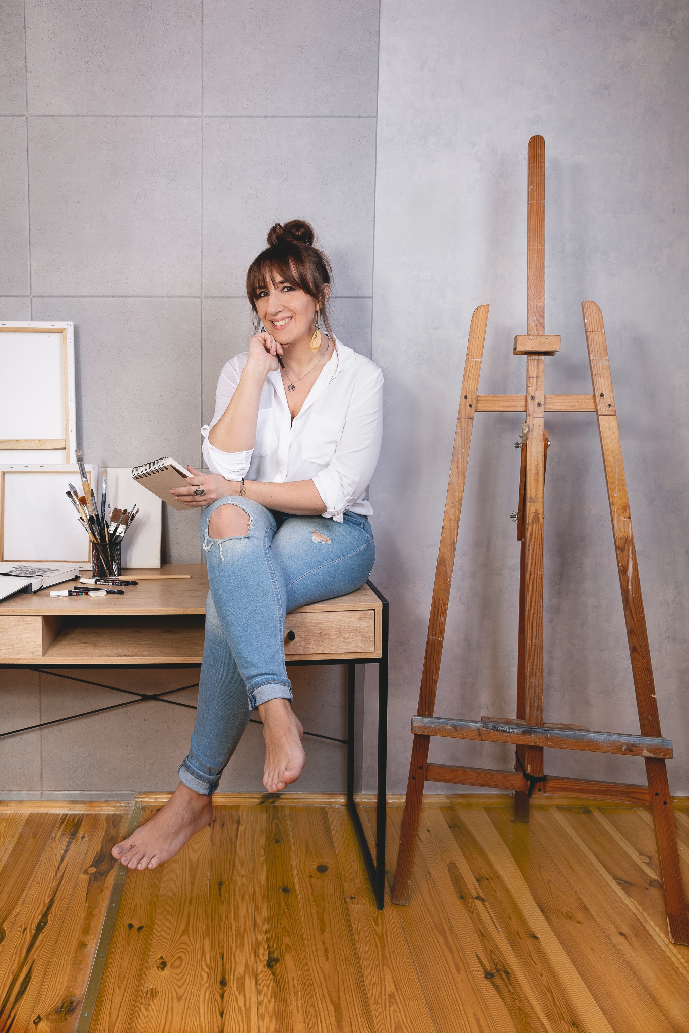 Uśmiechnięta kobieta siedzi na biurku z przyborami malarskimi obok drewnianego sztalugi w pracowni artystycznej z drewnianą podłogą i szarą ścianą.