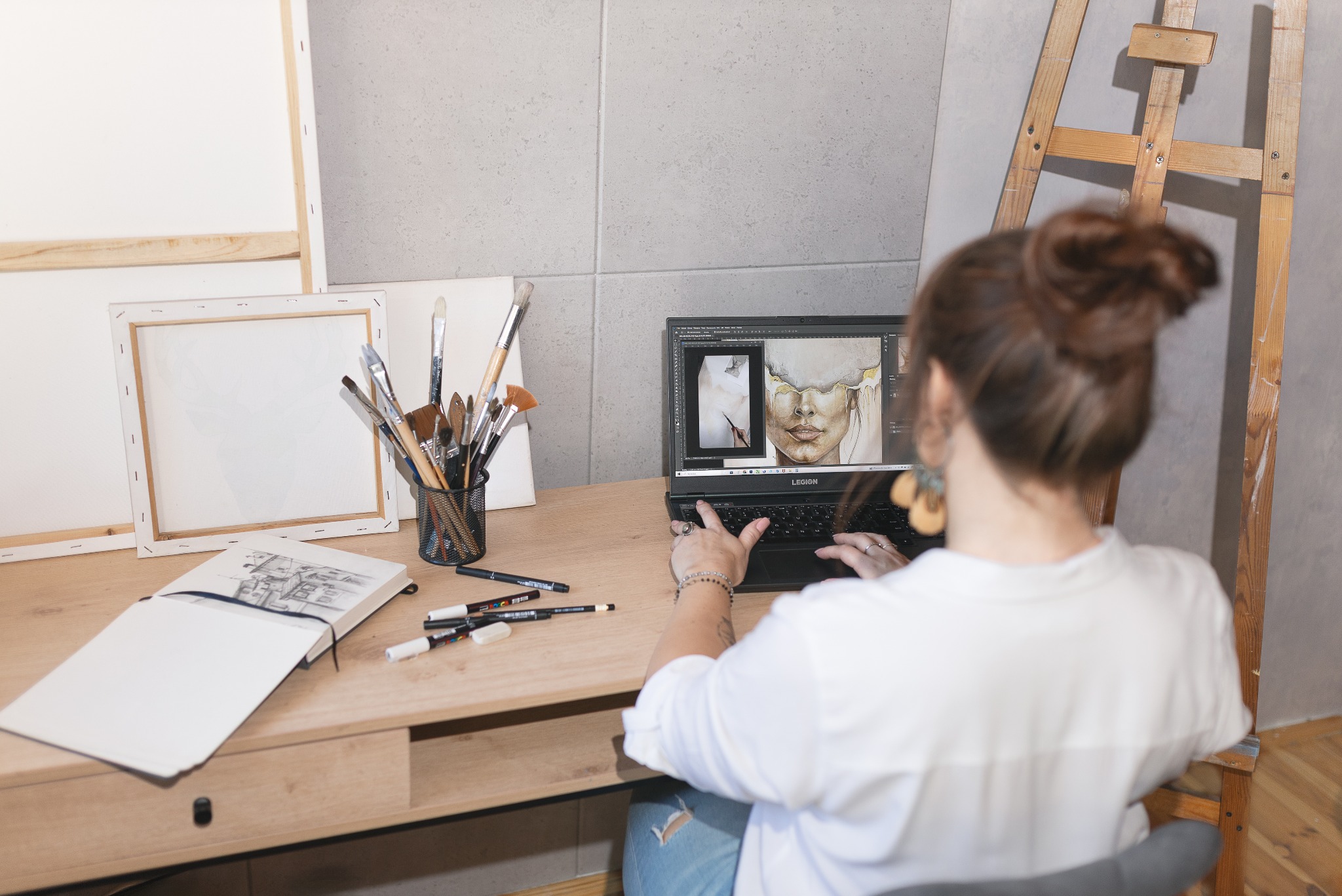 Artystka pracuje nad cyfrowym portretem na laptopie, otoczona pędzlami, szkicownikiem i płótnami. Widoczny drewniany statyw malarski i jasne, naturalne światło.