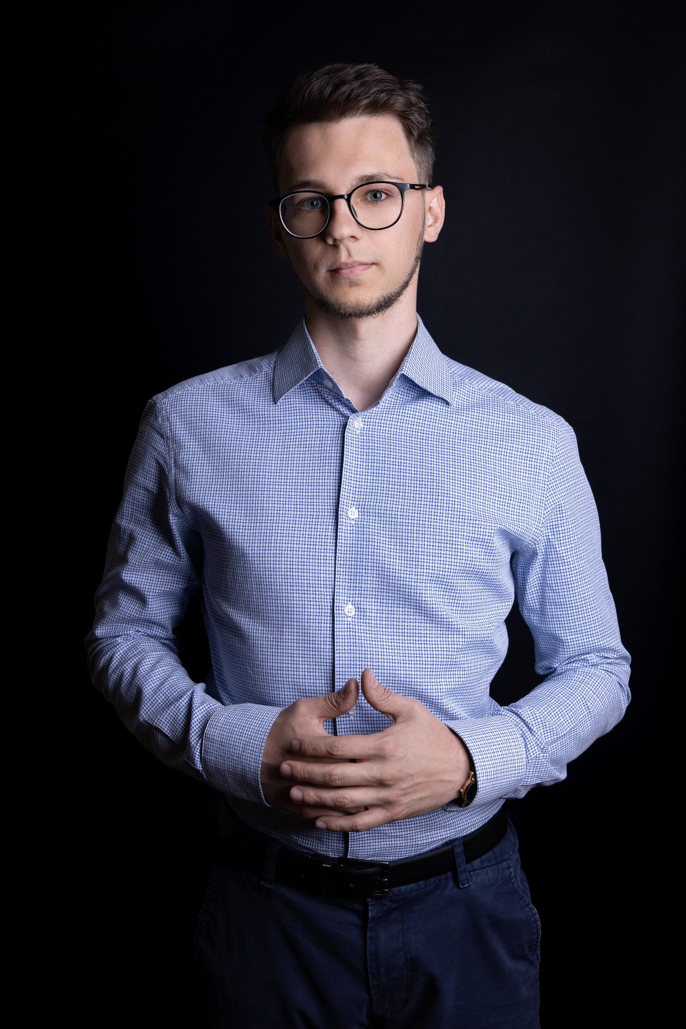 Mężczyzna w okularach i błękitnej koszuli na ciemnym tle, dłonie splecione na wysokości brzucha. Portret biznesowy.