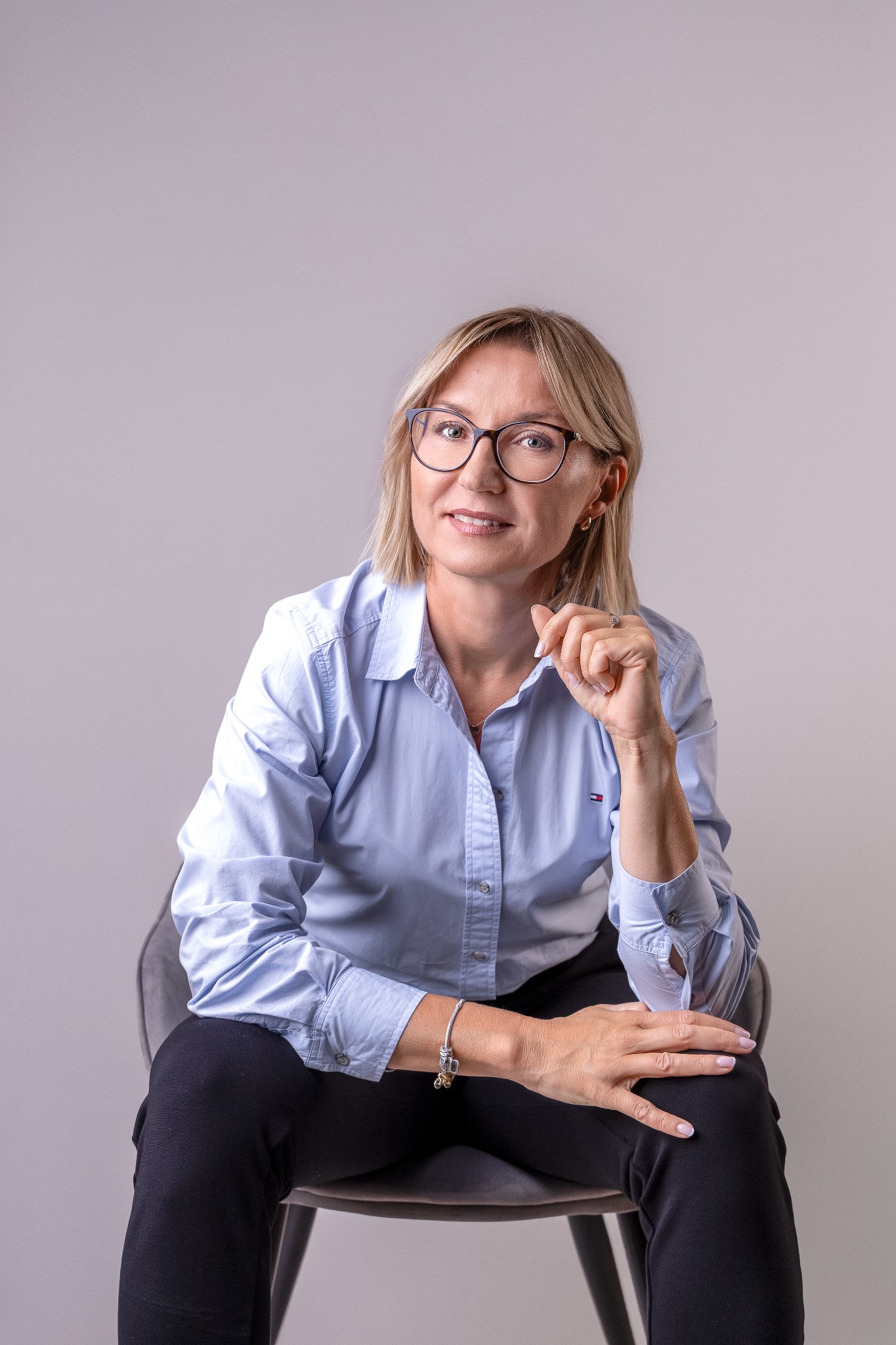 Portret kobiety w okularach, siedzącej na krześle, w błękitnej koszuli i czarnych spodniach. Uśmiechnięta, z dłonią przy twarzy. Stonowane tło, naturalne światło.