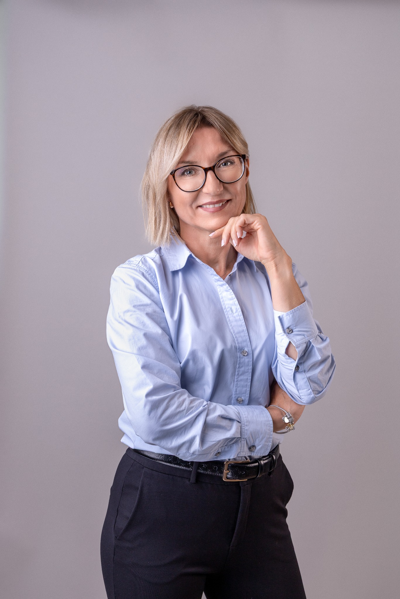 Portret uśmiechniętej kobiety w okularach, błękitnej koszuli i czarnych spodniach, z ręką podpartą pod brodą na neutralnym tle. Profesjonalny wizerunek.