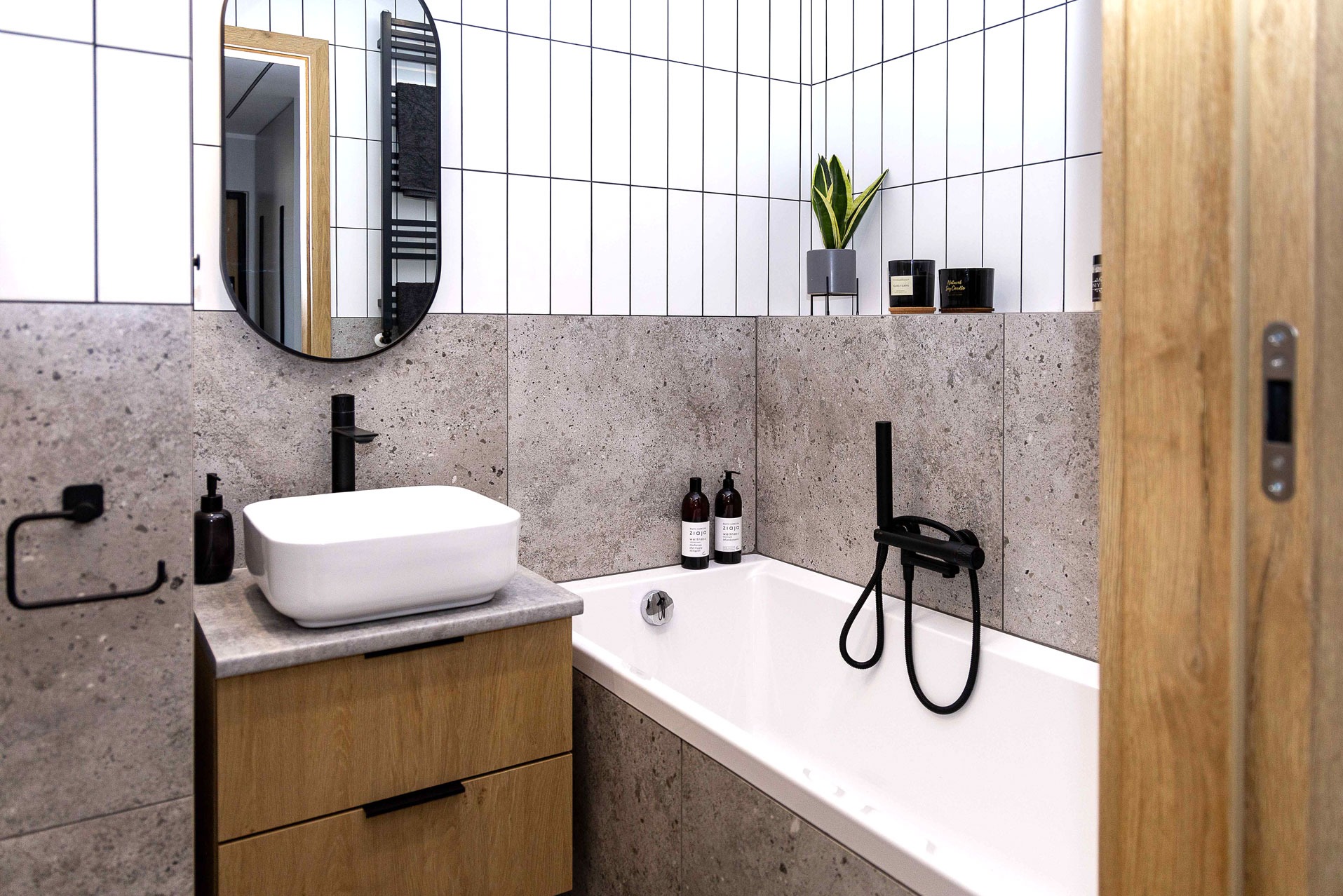 Nowoczesna łazienka z białą umywalką nablatową, wanną i czarną armaturą na tle szarych płytek imitujących kamień i białych, wąskich płytek na ścianie.