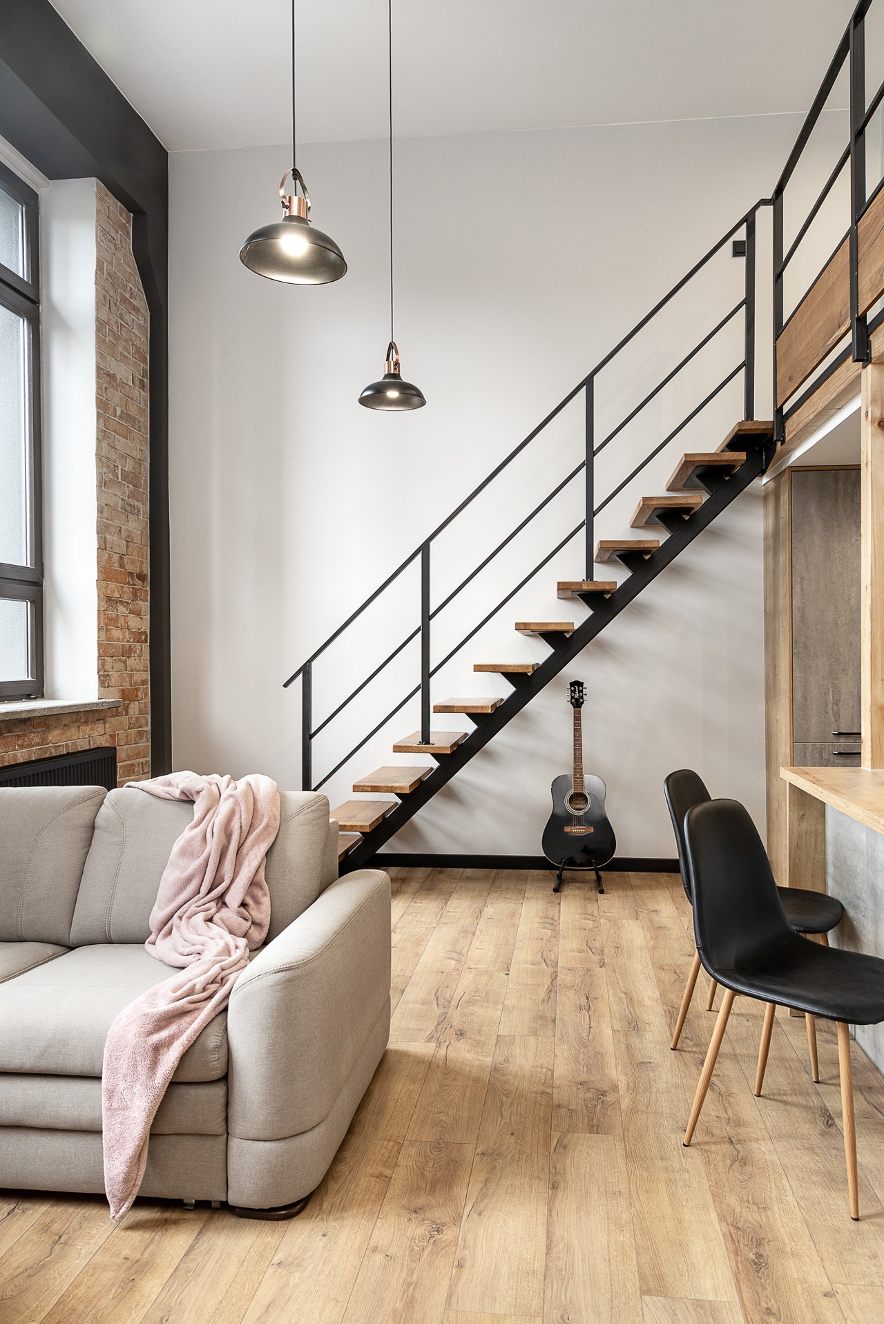 Jasne wnętrze loftu z drewnianymi schodami, szarą sofą z różowym pledem i czarną gitarą pod schodami. Dwie lampy wiszące nad podłogą z paneli.