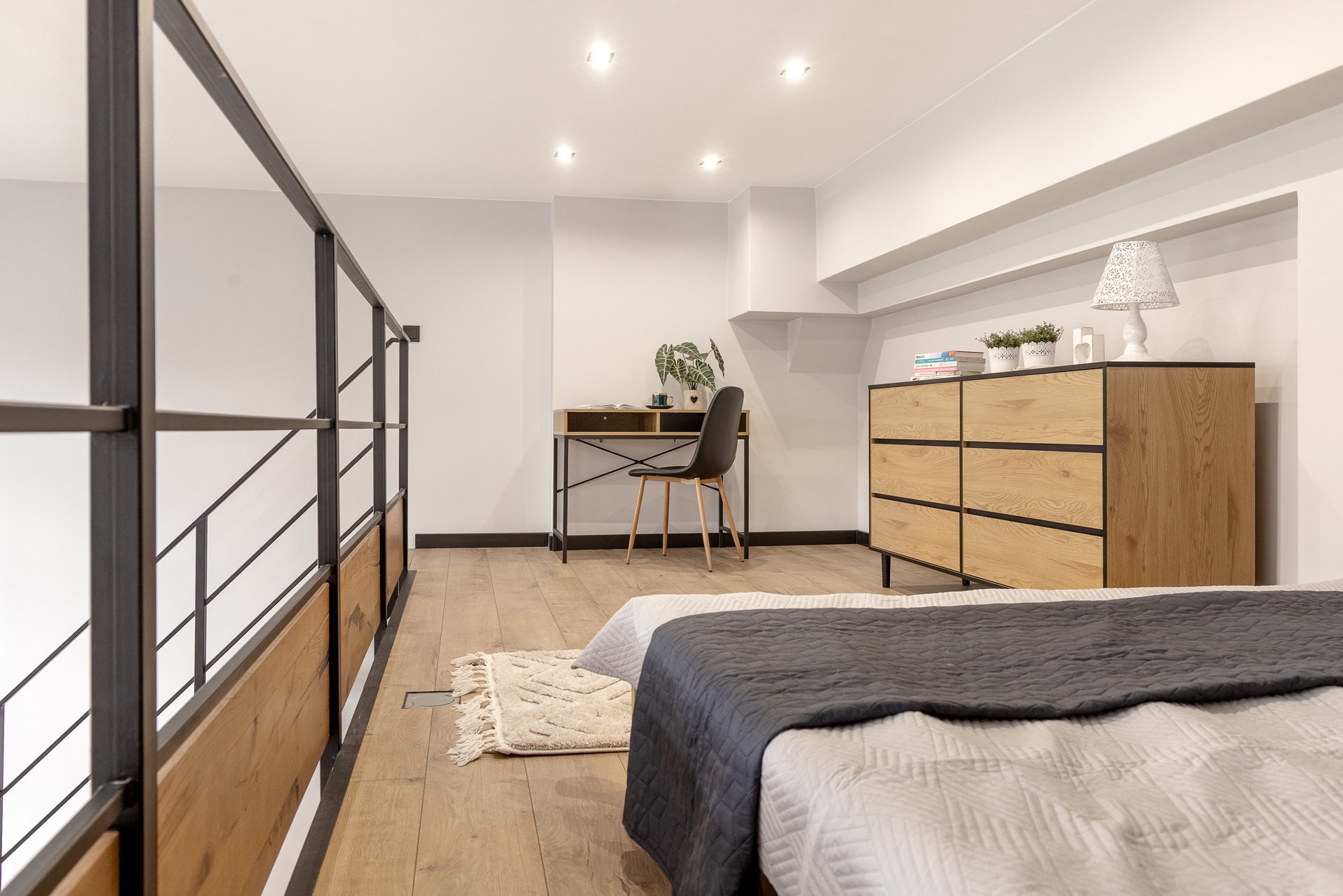 Jasne wnętrze z antresolą: drewniana podłoga, biurko z krzesłem, komoda z dekoracjami, fragment łóżka. Minimalistyczny design, neutralna kolorystyka, dobre oświetlenie.