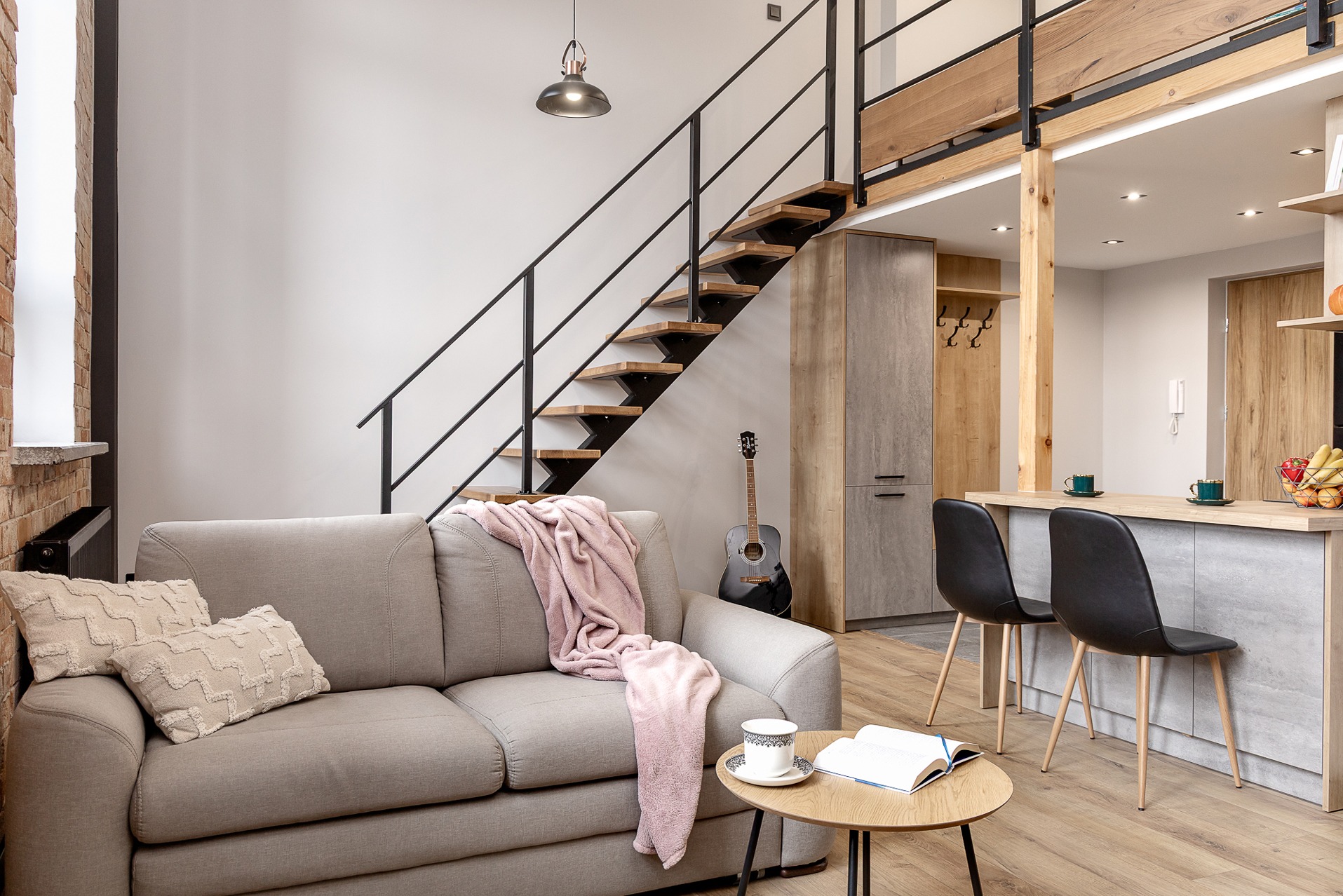 Stylowe wnętrze mieszkania z antresolą: szara sofa z poduszkami i różowym pledem, drewniane schody z czarną balustradą, gitara akustyczna w tle, nowoczesna kuchnia.