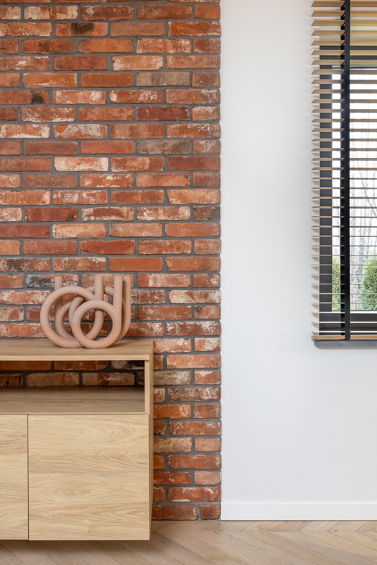 Minimalistyczny salon: ceglana ściana, drewniana komoda z dekoracją, żaluzje w oknie. Jasne wnętrze z parkietem w jodełkę.