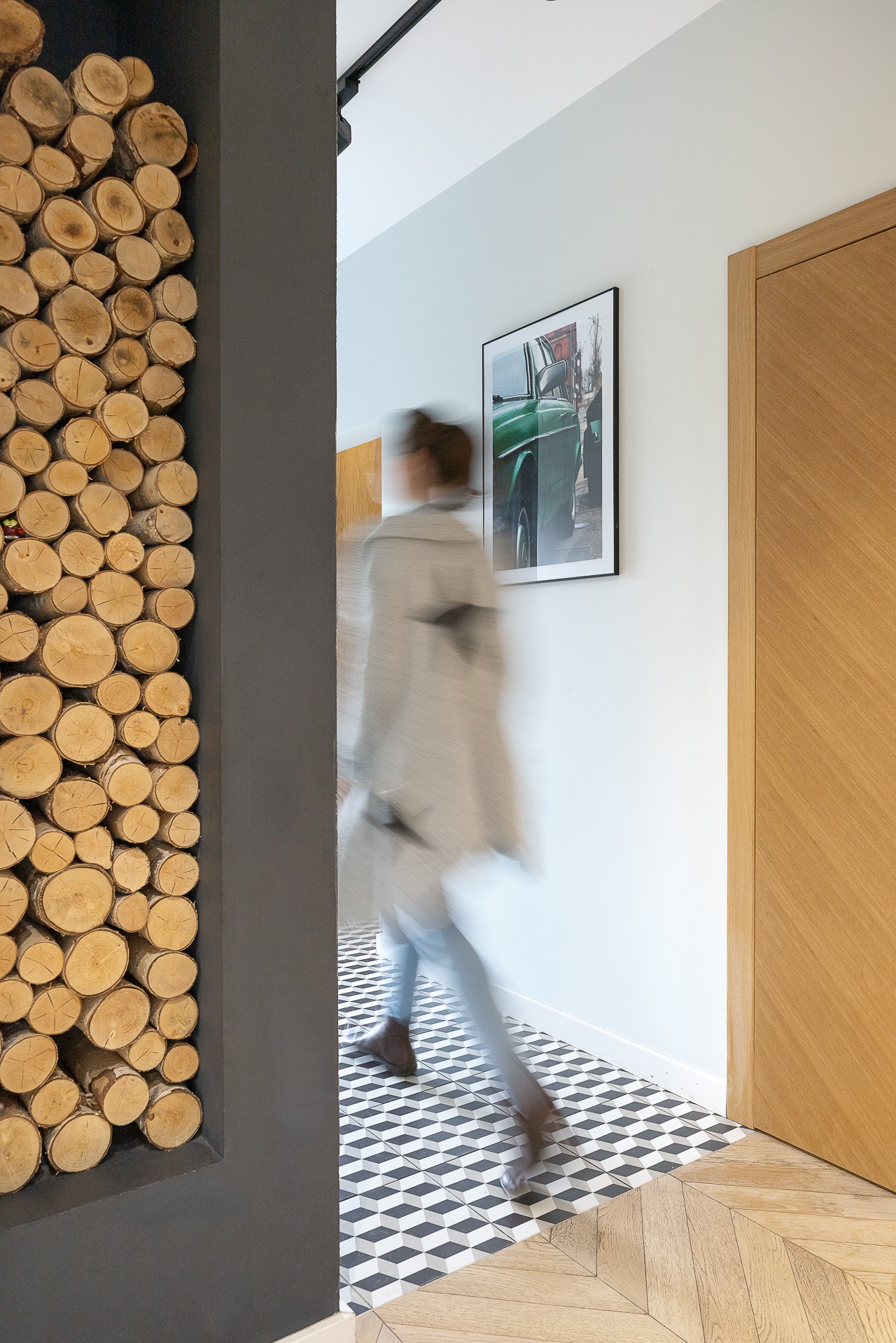 Nowoczesny hol z dekoracyjną ścianą z drewna, wzorzystą podłogą i obrazem zielonego samochodu. Rozmyta sylwetka osoby w ruchu dodaje dynamiki wnętrzu.