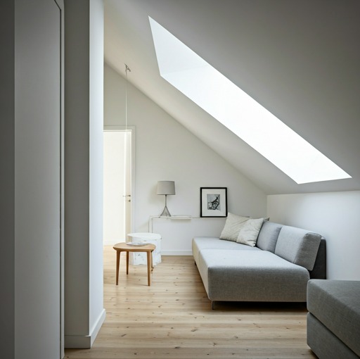 Jasne poddasze ze skośnym oknem dachowym, szarą sofą, drewnianą podłogą i minimalistycznym wystrojem. Widoczne świeżo pomalowane ściany na biało.