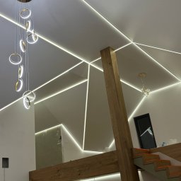 VIP GROUP TRADE SPÓŁKA Z OGRANICZONĄ ODPOWIEDZIALNOŚCIĄ - Nowoczesny salon z białym sufitem, geometrycznym oświetleniem LED, drewnianą konstrukcją i designerską lampą wiszącą z okrągłymi elementami.