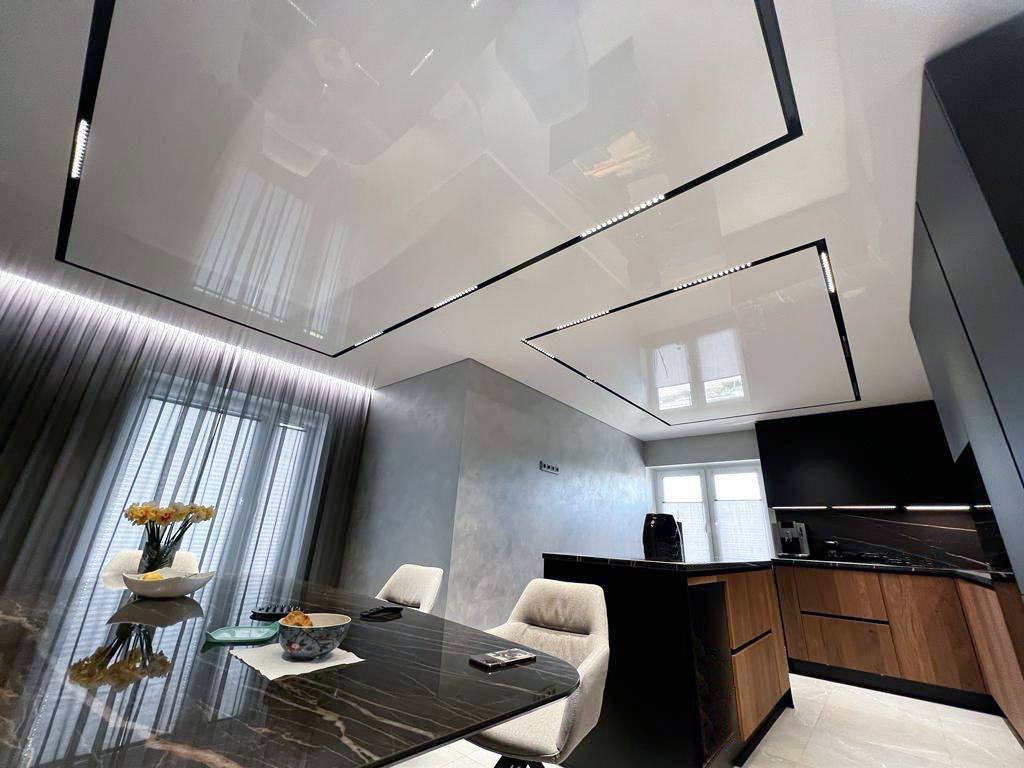 Biały, błyszczący sufit z czarnymi obramowaniami i oświetleniem LED w nowoczesnej jadalni z marmurowym stołem i częściowo widoczną kuchnią.