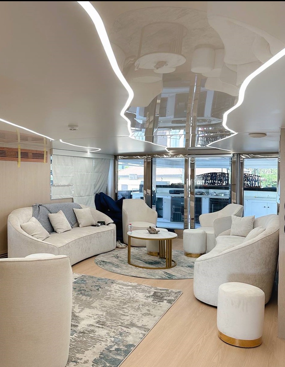 Nowoczesny salon z błyszczącym sufitem podwieszanym z liniowym oświetleniem LED, odbijającym wnętrze pokoju, w jasnej kolorystyce z meblami tapicerowanymi i drewnianą podłogą.