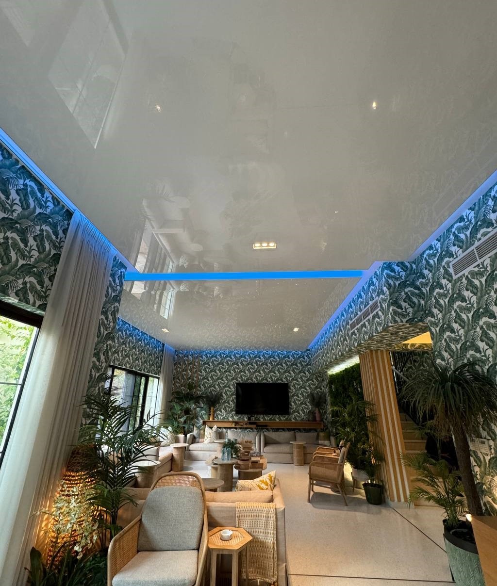 Biały, błyszczący sufit z niebieskim oświetleniem LED w nowoczesnym salonie z tapetą w tropikalne liście, widok z dołu.