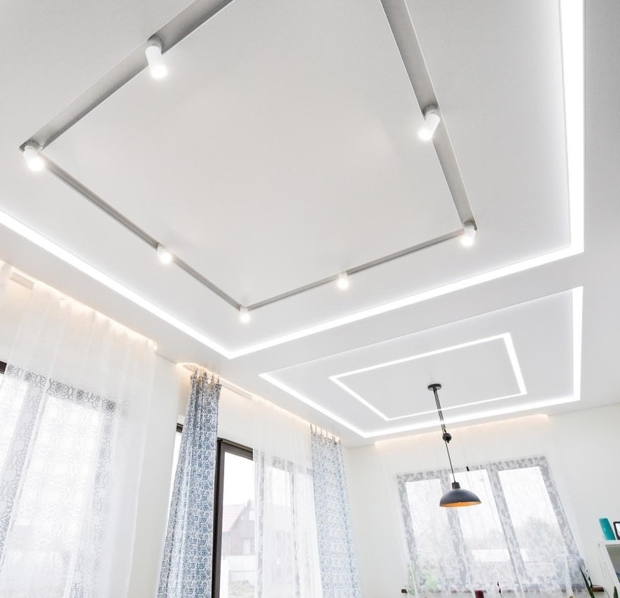 Wielopoziomowy biały sufit z oświetleniem LED i szynoprzewodami z reflektorami punktowymi, widok z dołu na okna z firanami.