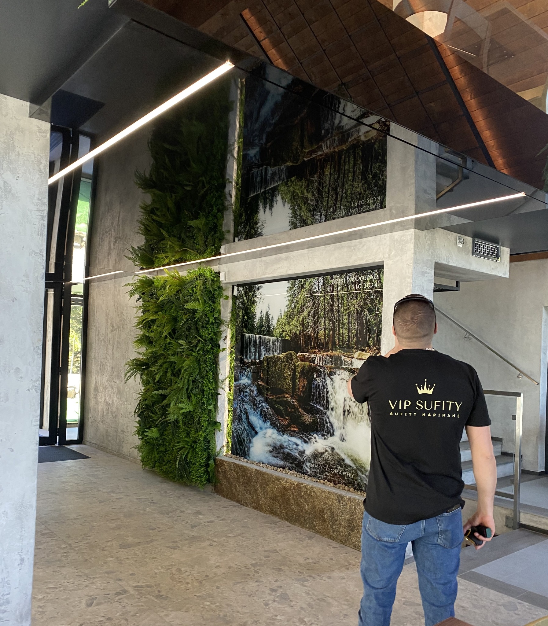 Nowoczesne wnętrze z podwieszanym sufitem lustrzanym, oświetleniem LED, pionową zielenią i fototapetą z motywem wodospadu. Osoba w firmowej koszulce.