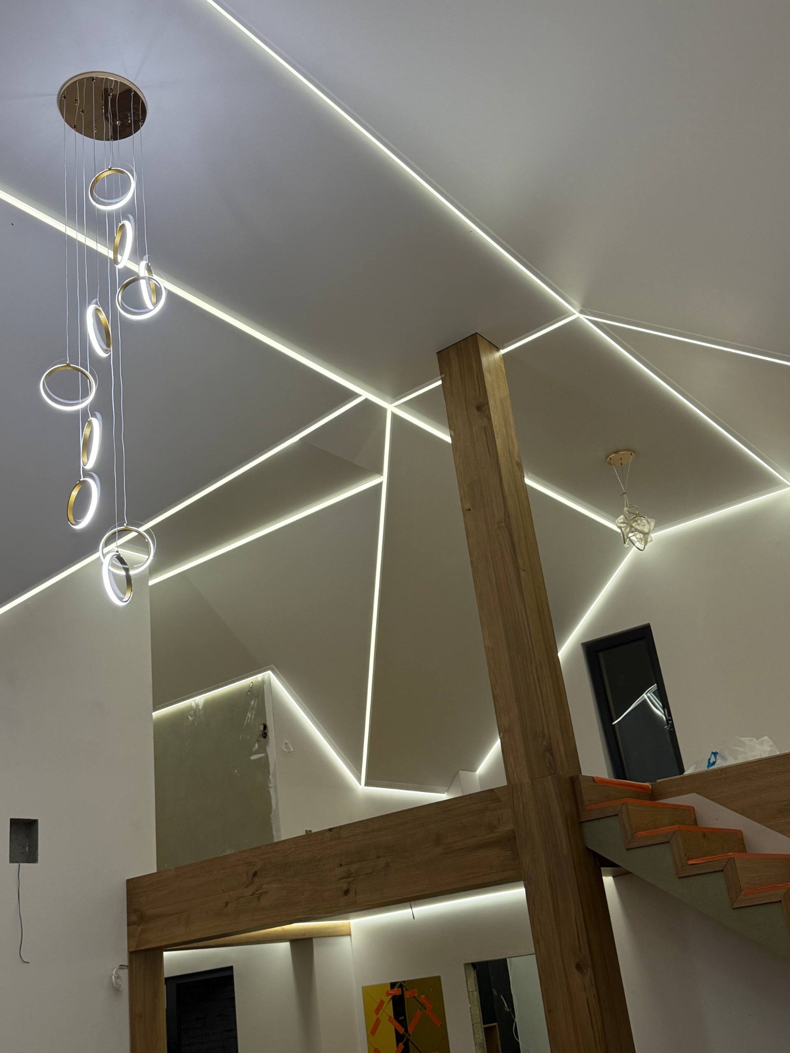 Nowoczesny salon z białym sufitem, geometrycznym oświetleniem LED, drewnianą konstrukcją i designerską lampą wiszącą z okrągłymi elementami.