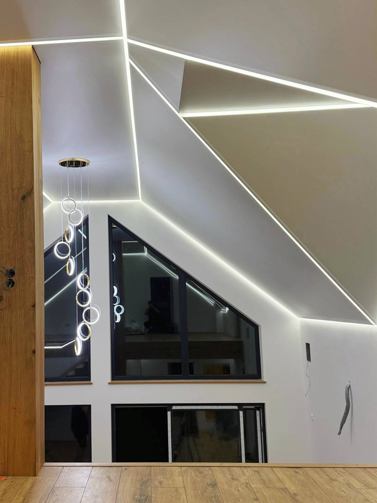 Nowoczesny sufit z oświetleniem LED w kształcie geometrycznym, wisząca lampa z okręgami, duże okno. Minimalistyczny design w jasnych barwach.