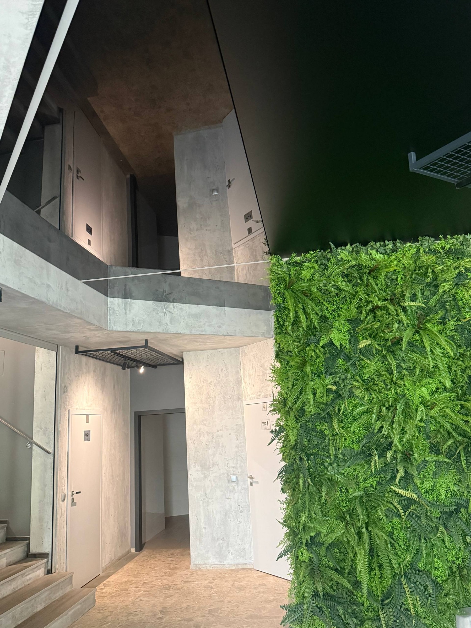 Nowoczesne wnętrze biurowe z zieloną ścianą i betonowym stropem. Widoczne schody, drzwi i oświetlenie. Minimalistyczny design z elementami natury.