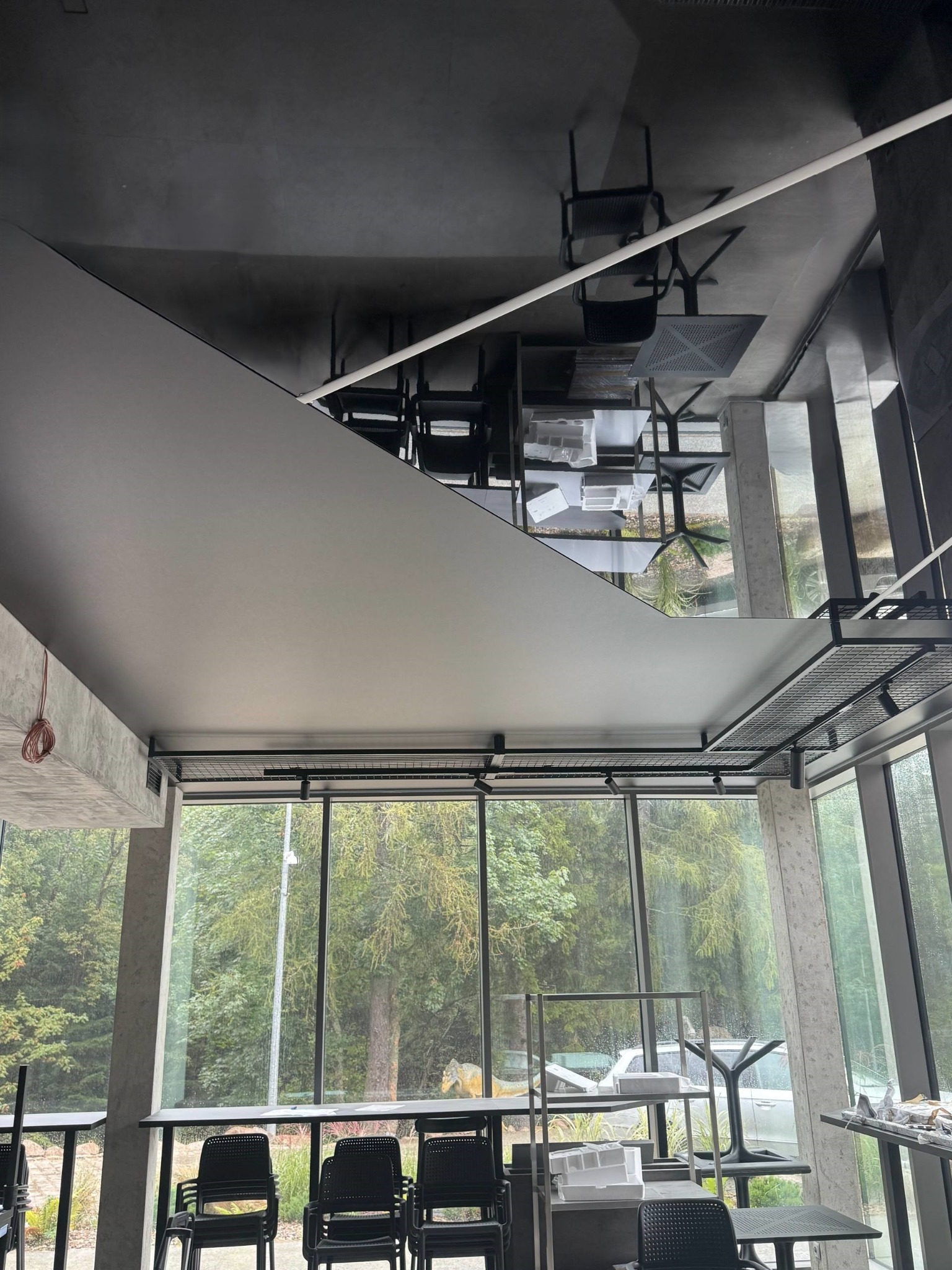 Nowoczesne wnętrze z błyszczącym, szarym sufitem i dużymi oknami z widokiem na zieleń. Widoczne elementy oświetlenia i odbicia w suficie.