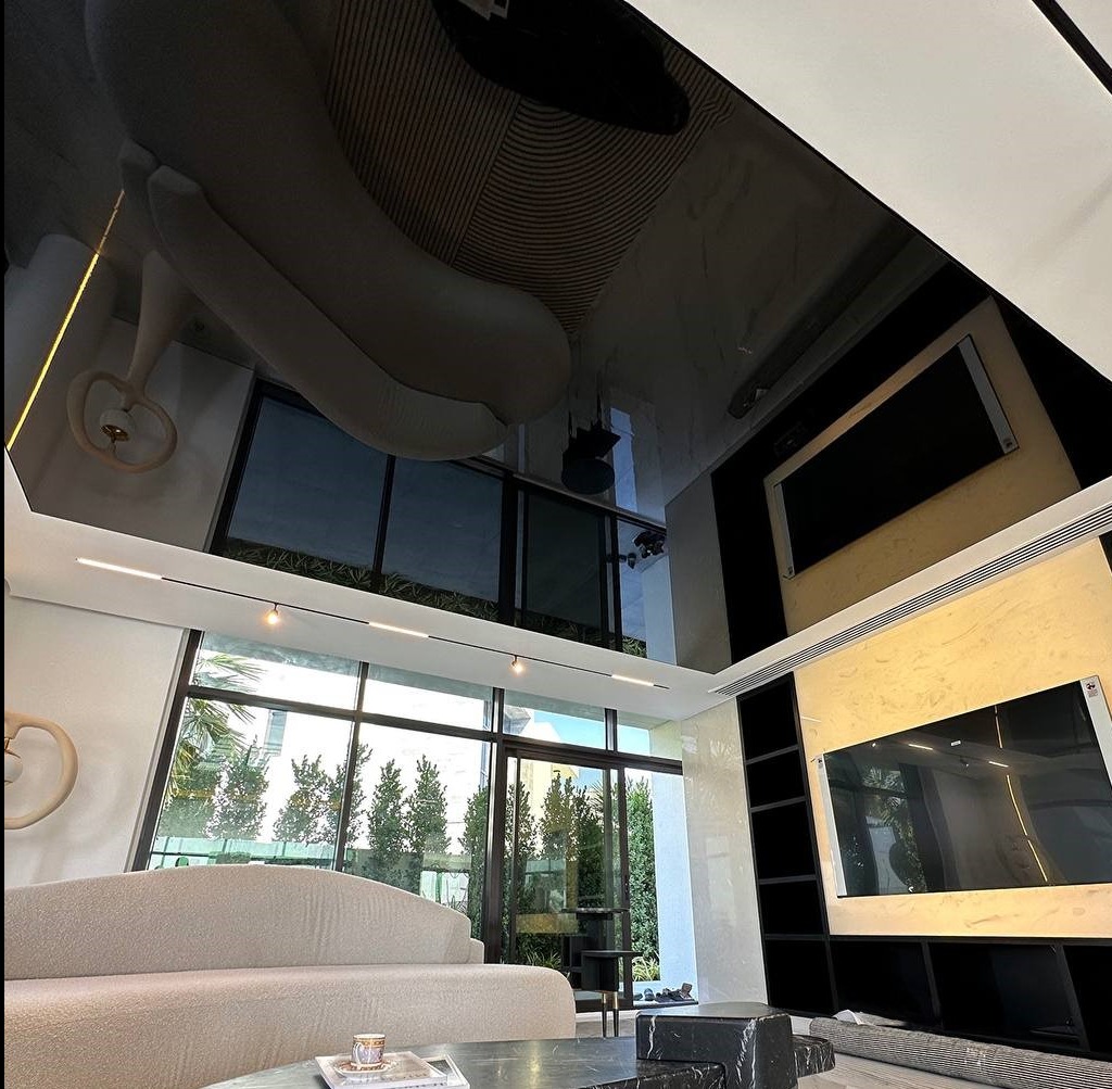 Salon z błyszczącym, czarnym wykończeniem sufitu, odbijającym wnętrze. Widoczne okna, sofa i elementy dekoracyjne. Oświetlenie liniowe podkreśla nowoczesny charakter.