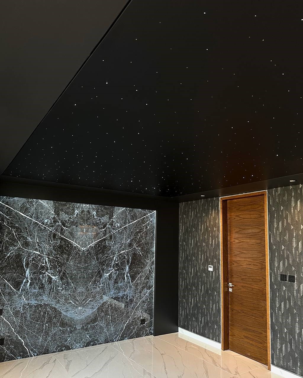 Nowoczesny pokój z czarnym sufitem imitującym gwiaździste niebo. Ściana z efektem marmuru i wzorzysta tapeta. Brązowe drzwi i jasna podłoga z płytek.