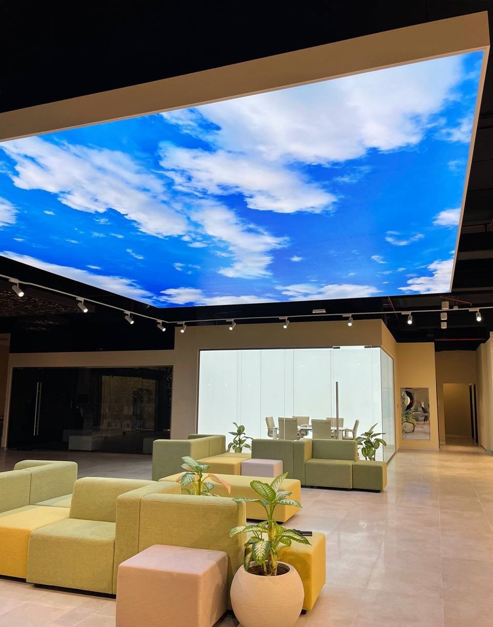 Nowoczesne wnętrze z podświetlanym sufitem imitującym niebo z chmurami, miękkie sofy w odcieniach zieleni i żółci, rośliny doniczkowe, przeszklona sala konferencyjna.