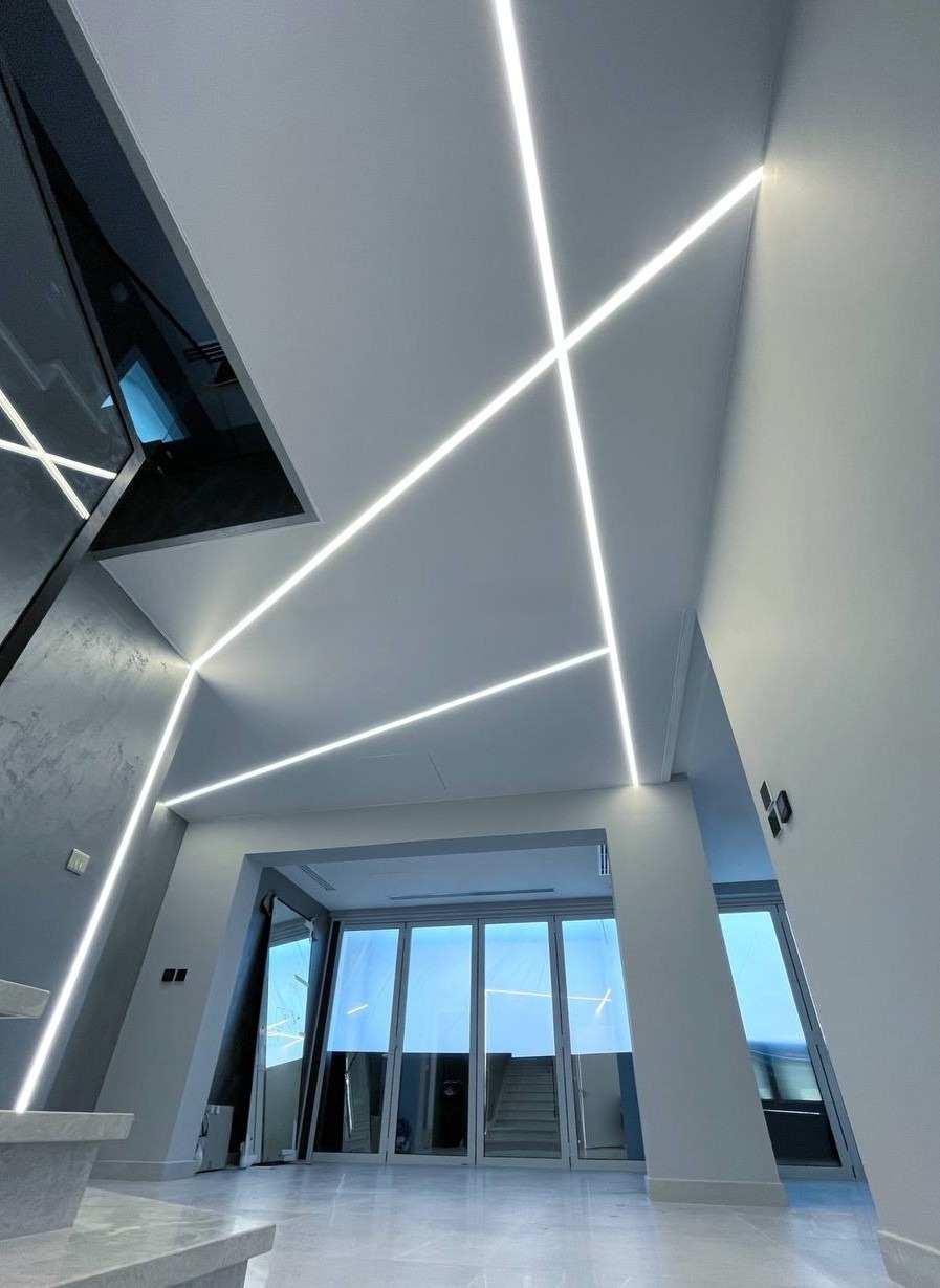 Nowoczesne wnętrze z geometrycznym oświetleniem LED na suficie i ścianach, tworzące unikalny wzór. Widoczne schody i przeszklone drzwi.