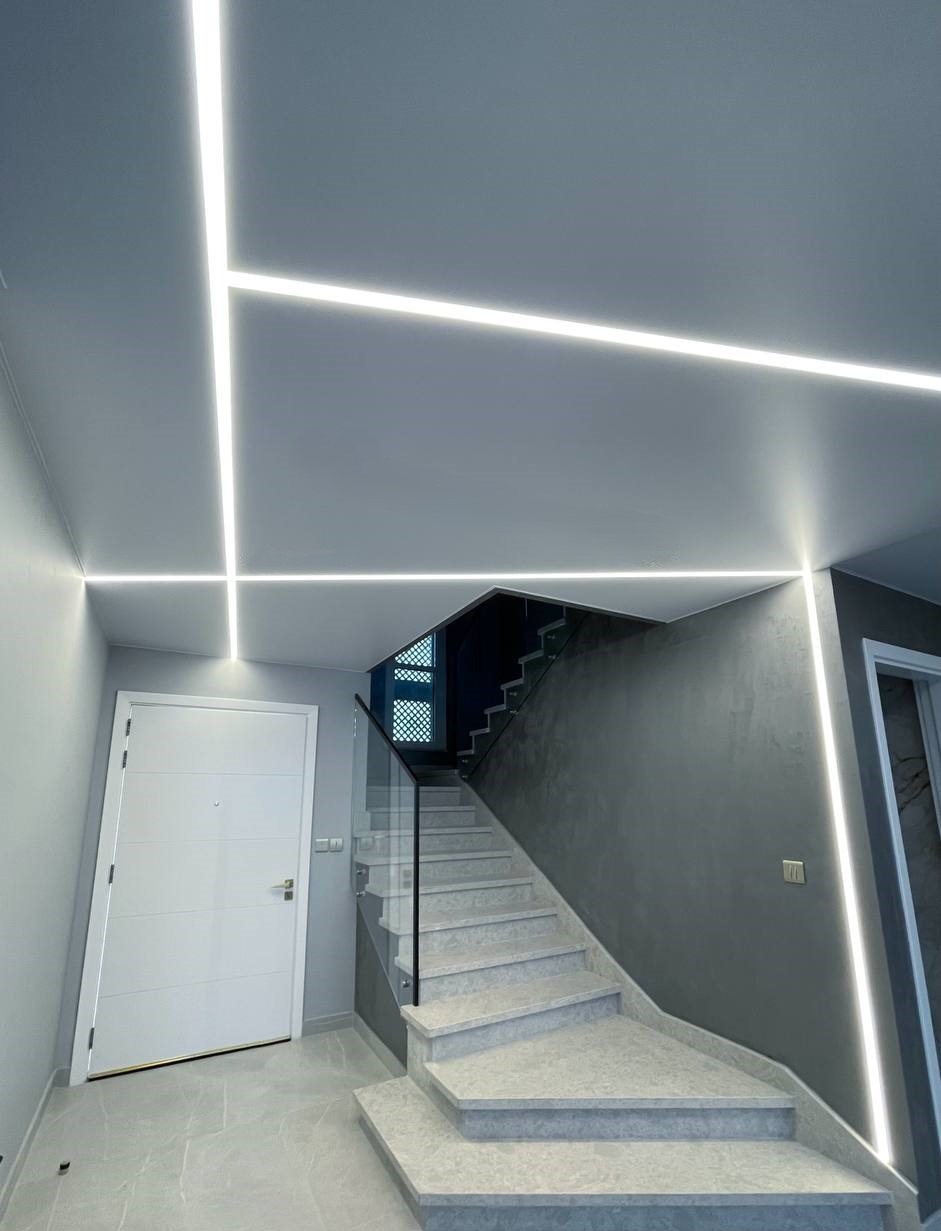 Nowoczesne oświetlenie LED na suficie i ścianie w korytarzu z schodami. Geometryczne linie światła podkreślają minimalistyczny design wnętrza.
