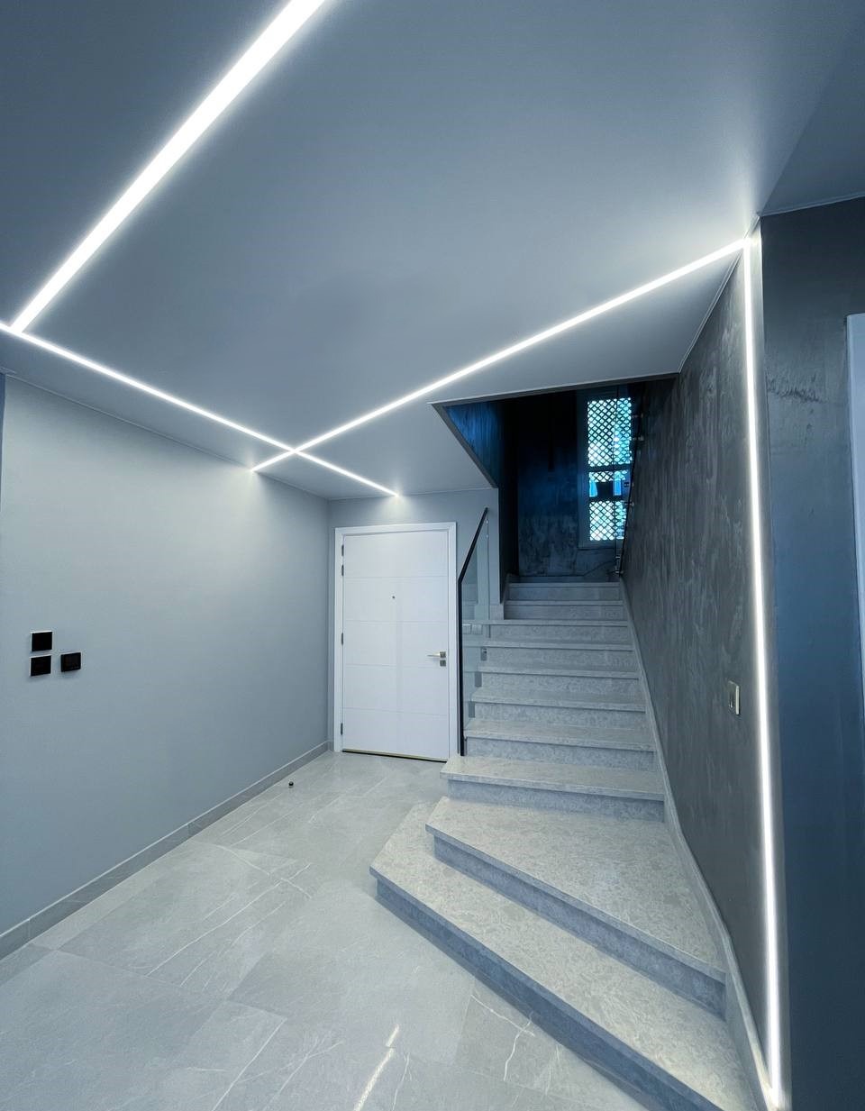 Nowoczesne oświetlenie LED w korytarzu z marmurowymi schodami i białą stolarką drzwiową. Minimalistyczny design z liniowymi lampami sufitowymi.