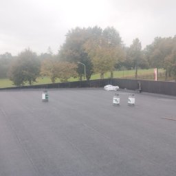 Diza Golden Company - Płaski, czarny dach pokryty papą z widocznymi zgrzewami, na którym stoją trzy butle z gazem i leży zwinięta papa, w tle drzewa i pochmurne niebo.