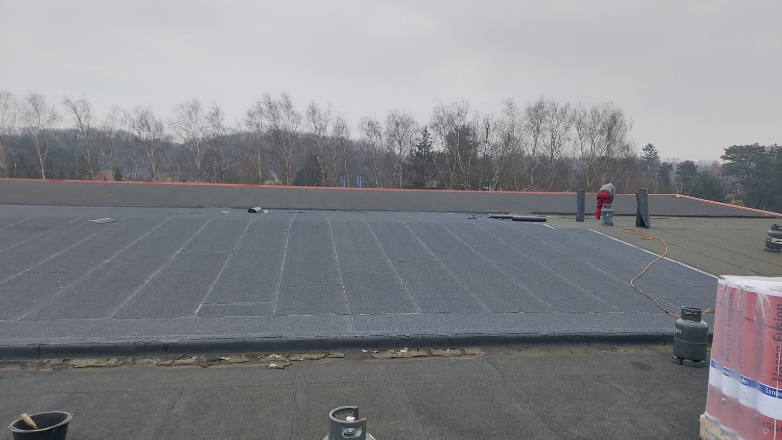 Prace na dachu płaskim: dekarz w czerwonych spodniach układa papę termozgrzewalną, widoczne butle z gazem i zgrzewarka, w tle nagie drzewa.