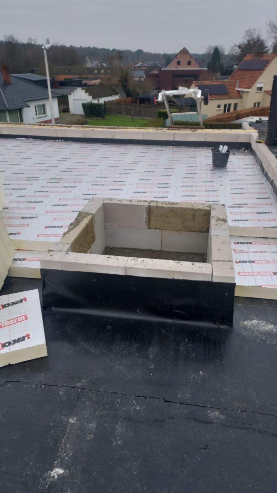 Płaski dach w trakcie budowy z wykończonym kominem wentylacyjnym, widoczne warstwy izolacyjne i papa dachowa.