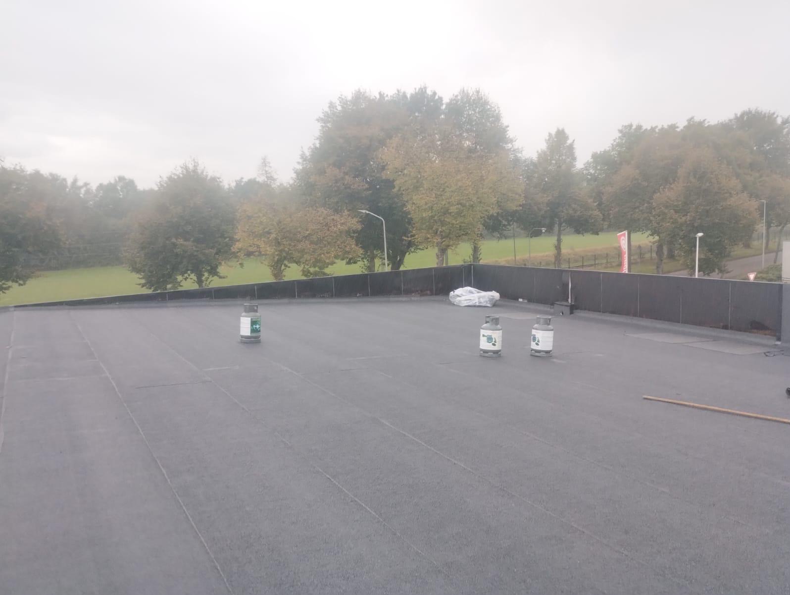 Płaski, czarny dach pokryty papą z widocznymi zgrzewami, na którym stoją trzy butle z gazem i leży zwinięta papa, w tle drzewa i pochmurne niebo.