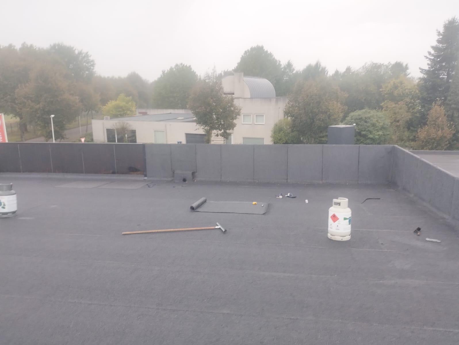 Bitumiczna papa na płaskim dachu budynku w trakcie prac dekarskich, widoczny gazowy palnik dekarski, rolka papy, oraz narzędzia dekarskie. W tle budynek i drzewa w lekkiej mgle.