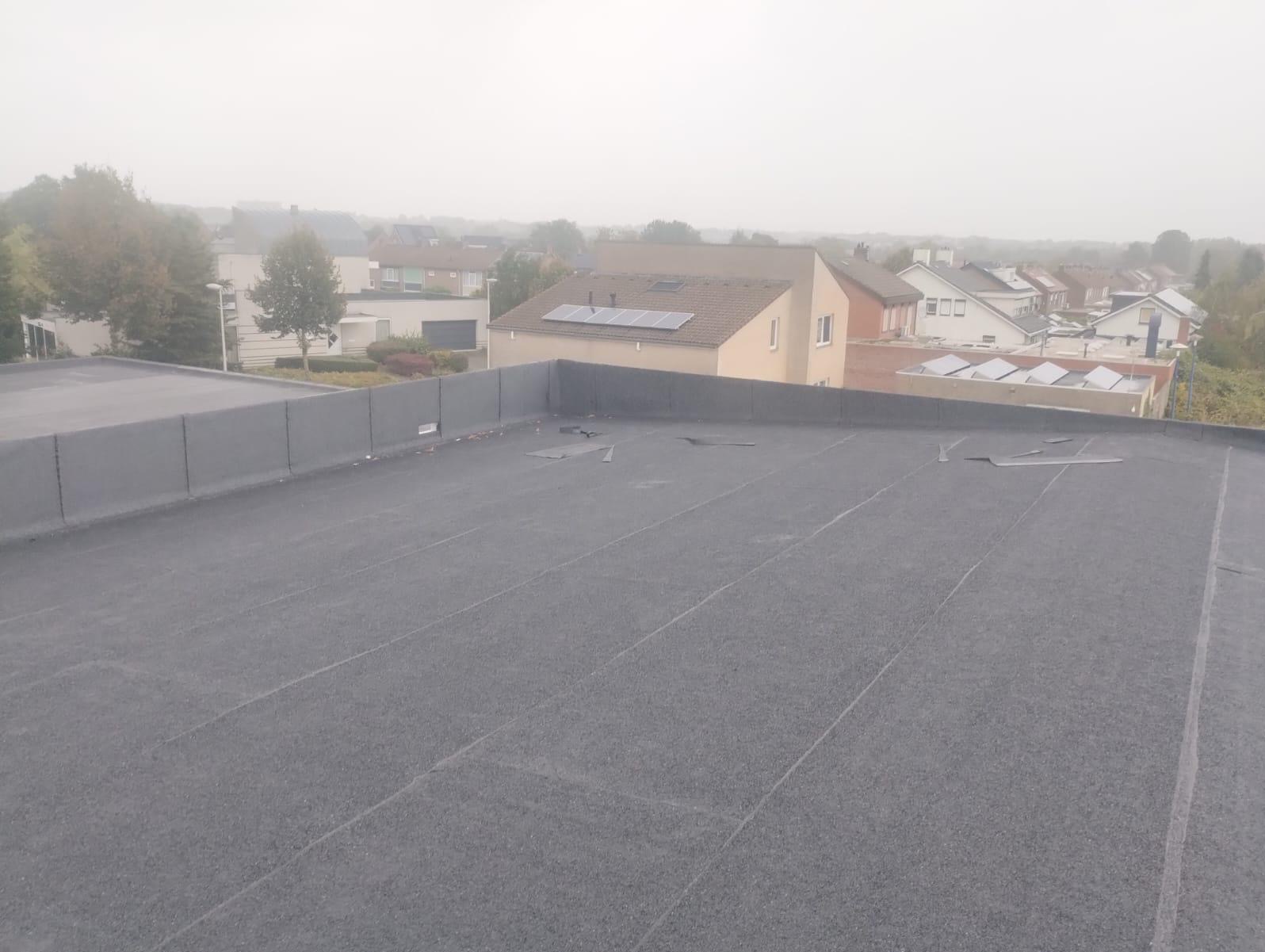 Widok z góry na płaski dach pokryty papą termozgrzewalną, z widocznymi łączeniami i fragmentami odciętej papy, na tle zabudowy jednorodzinnej w mglisty dzień.