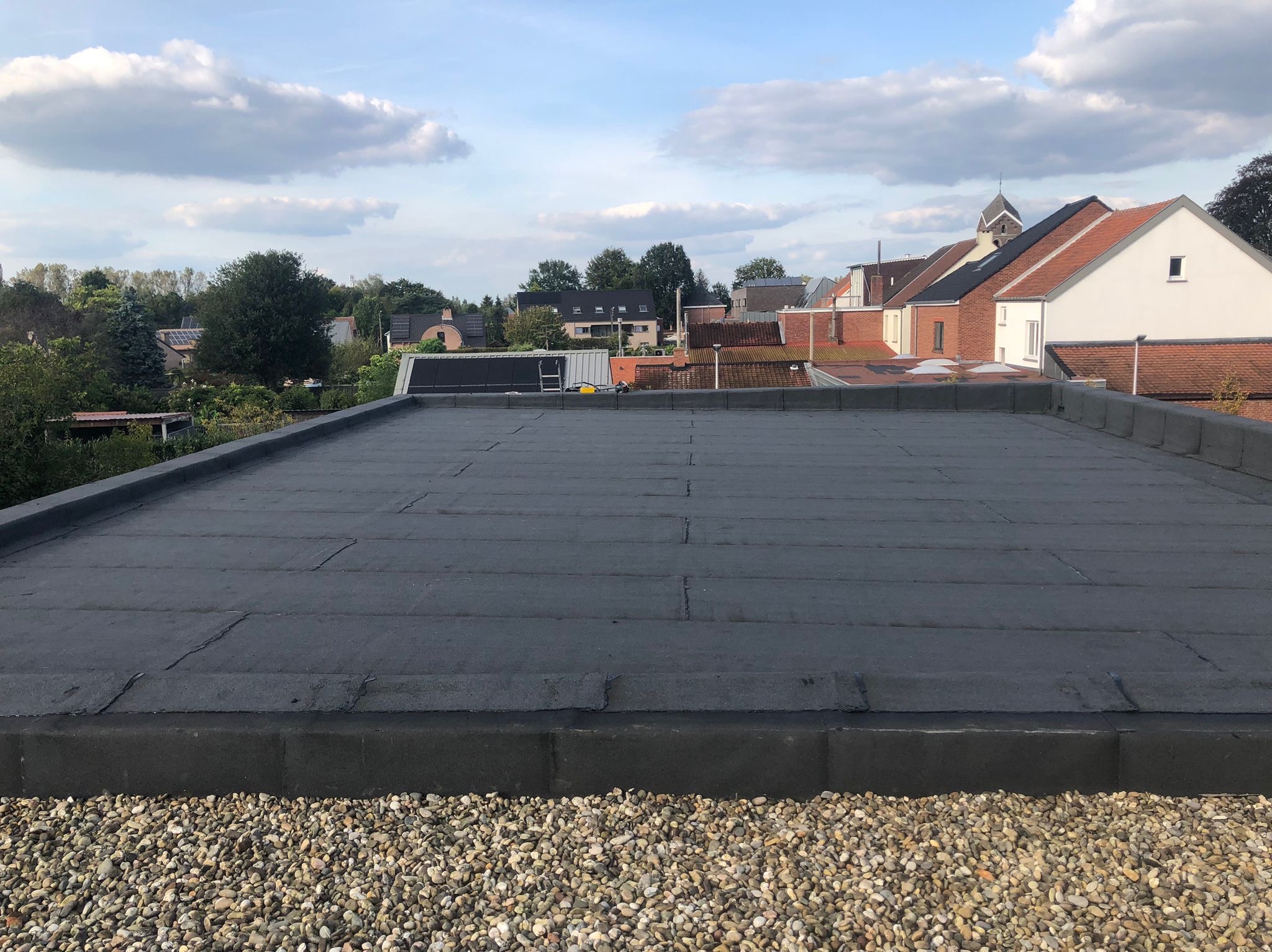 Widok z góry na płaski dach pokryty papą, z widocznymi łączeniami i obrzeżem, w tle zabudowa jednorodzinna i błękitne niebo z chmurami.