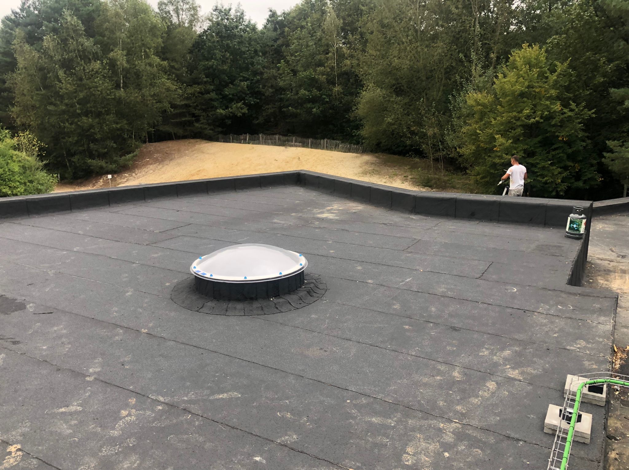 Płaski dach pokryty czarną papą z zamontowanym świetlikiem kopułkowym; w tle widoczny pracownik na drabinie oraz zalesiony teren.