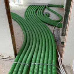 ECO-CIEPEŁKO SPÓŁKA Z OGRANICZONĄ ODPOWIEDZIALNOŚCIĄ - Zielone rury instalacji ogrzewania podłogowego, ułożone równolegle na surowej posadzce, widoczne metalowe mocowania i fragmenty instalacji wentylacyjnej pod sufitem.
