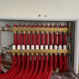 ECO-CIEPEŁKO SPÓŁKA Z OGRANICZONĄ ODPOWIEDZIALNOŚCIĄ - Rozdzielacz ogrzewania podłogowego z czerwonymi rurami w szafce, widoczne manometry i białe regulatory przepływu, ułożony na macie grzewczej.